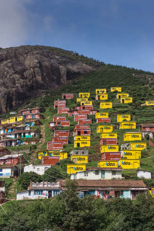Ooty, India - October 25, 2013: Valparai village looks like a favela built on the slope of a hill. Bright yellow and wine red houses for poor people sponsored and painted by Idea and Vodaphone. Blue sky, rocky hill, green foliage, people.の写真素材