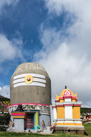 Ooty, India - October 25, 2013: Shiva shrine near Valparai village in the form of giant Shivalingam on top of circular construction. Blue and white cloudy sky. Houses in far background on hill. Statues on shrine.のeditorial素材