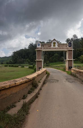 Ooty, India - October 25, 2013: Small rectangular side gate over narrow road into Wellington Cantonment. Features names and logos of garrison and college. Stormy sky, green foliage of forest in back. Hill and meadows.のeditorial素材