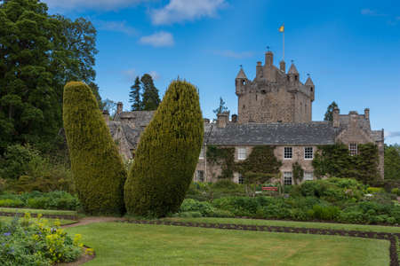 Cowdor, Scotland - June 2, 2012: The brown-gray stone structure of undamaged Cowdor Castle under blue sky. Flag on top. Large part of formal garden in forefront. Green foliage.のeditorial素材