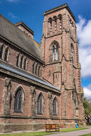 Inverness, Scotland - June 1, 2012: Shot of one flat tower and the side of brown stone Saint Andrews Cathedral under blue sky and white clouds. Brown bench on green lawn.の写真素材