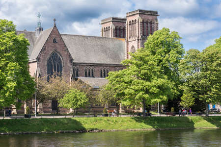 Inverness, Scotland - June 1, 2012: Brown stone Saint Andrews Cathedral show side, roof and the two towers. White cloudy sky. Seen from across Ness River. Green trees.のeditorial素材