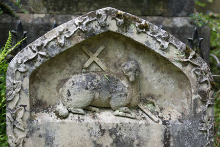 Grasmere, England - May 30, 2012: In the cemetery of the Saint Oswald Church stands the gray and white old tombstone showing the Agnus Dei, the Lamb of God with a cross. Green foliage.のeditorial素材