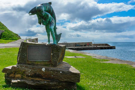 Dunbeath, Scotland - June 4, 2012: Kenn and the Salmon statue stands in the harbor. Green meadow, part of the harbor pier, blue sky with large clouds. Scene out of Highland River book written by Neil Gunn.のeditorial素材
