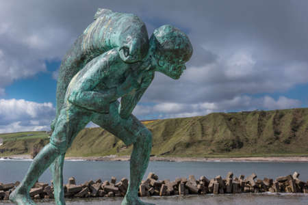 Dunbeath, Scotland - June 4, 2012: Closeup of Kenn and the Salmon statue which stands in the harbor. Green cliffs and North Sea in background. Dark cloudy sky with blue patch. Scene out of Highland River book written by Neil Gunn.のeditorial素材