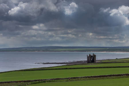 Nybster, Scotland - June 4, 2012: Wide long shot of on the shore of a calm North Sea at the bay stands the ruin of the local strong house or castle under cloudy sky. Green meadows in front. Darker hills and beach on other side of bay.のeditorial素材