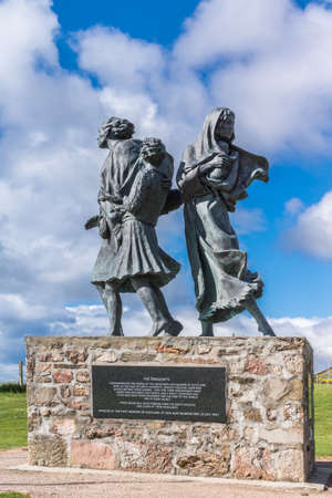 Helmsdale, Scotland - June 4, 2012: The Emigrant Statue at the south entrance of the town. Lateral view on the four figures. Set on green hill. Blue sky with clouds, bronze with green spots. Dedication plaque.のeditorial素材