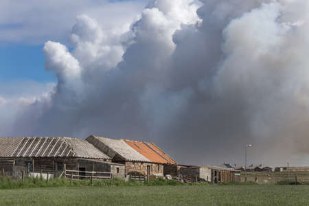 John O Groats, Scotland - June 4, 2012: Towering dark smoke clouds of large wildfire near John O Groats village threatens farm buildings. Green meadows, gray and red buildings. One blue sky patch.のeditorial素材
