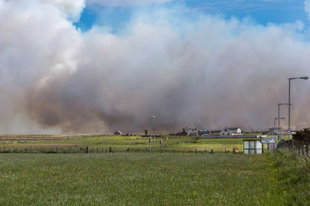 Towering dark smoke clouds of large wildfire near John O Groats village threatens farm buildings. Green meadows, gray and red buildings. One blue sky patch.のeditorial素材