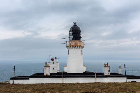 The lighthouse complex and tower at Dunnet Head against light blue sky. White towers with golden rim and black lighthouse cap with antennas. Set on brown grassy field.の写真素材
