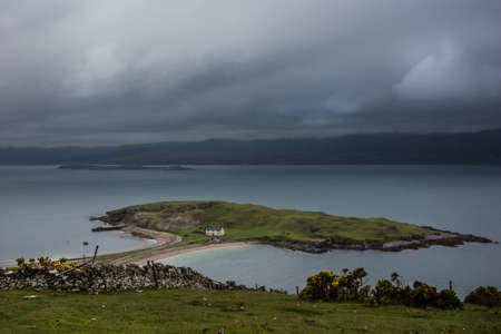 North Coast, Scotland - June 6, 2012: Small green island in Loch Eriboll, connected with a short dam, and featuring a white fisherman house. Cloudy foggy sky, bluish water, mountainous background.のeditorial素材