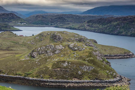 Aerial view over green rocky land tongue protruding into Loch Inchard blue waters. Horizon of mountains and dark clouds.の写真素材
