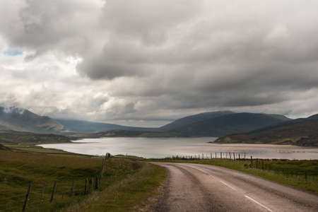 The rural road called highway descends on Kyle of Durness, part of Atlantic Ocean. Heavy dark and...