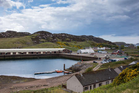 Northwest Coast, Scotland - June 6, 2012: The industrial and fishing section of Scourie harbor shows the docks, warehouses machines and more. Against gray rocky hills with green pasture patches under blue and white sky. Yellow broom flowers.のeditorial素材