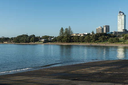 Auckland, New Zealand - March 2, 2017: Black volcanic south shore of Tapapuna Beach shows park, highrise and canoe rowers under blue sky with reflecting sea water.のeditorial素材