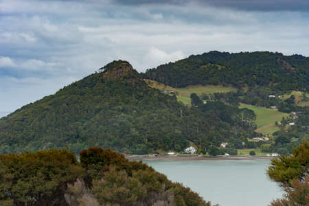Auckland, New Zealand - March 2, 2017: Forested Whatipu mountain at entrance of Huia Bay under white clouds in blue sky. Dwellings, foliage, pasture and blue-green water.のeditorial素材