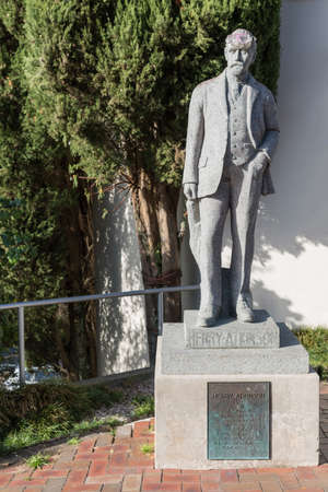 Auckland, New Zealand - March 2, 2017: Gray stone statue of Henry Atkinson, environmentalist, standing on stone cube in the village of Titirangi, Waitakere Ranges. Green foliage. Goggles on head.のeditorial素材
