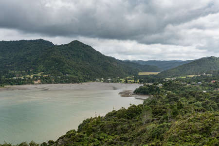 Auckland, New Zealand - March 2, 2017: Deep end of Huia Bay with mud flats and mountains under heavy cloudscape. Green foliage, green-gray waters, hidden dwellings.のeditorial素材