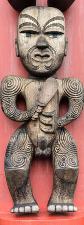 Auckland, New Zealand - March 2, 2017: Closeup of Te Hawiti Ancestor wood sculpture on totem pole in front of Arataki Maori Heritage Center. Brown wood of naked man on scarlet background.のeditorial素材