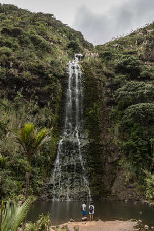 Auckland, New Zealand - March 2, 2017: Silver Karekare waterfall streams between green forested cliffs into dark pool. Two people at bottom. Gray skies.のeditorial素材