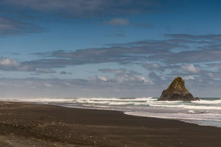 Auckland, New Zealand - March 2, 2017: Black volcanic sand Karakare Beach under blue cloudy sky with, Te Kaka Whakaara Rock (Watchman) in the surf of Tasman Sea.のeditorial素材