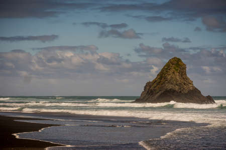 Auckland, New Zealand - March 2, 2017: Black sand Karakare Beach under blue cloudy sky with, Te Kaka Whakaara Rock (Watchman) in the surf of Tasman Sea.のeditorial素材