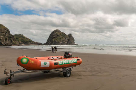 Auckland, New Zealand - March 2, 2017:  Closeup of orange dinghy boat of beach life guard at Piha Beach with Rabbit rock in the back. Tasman sea surf and people.のeditorial素材