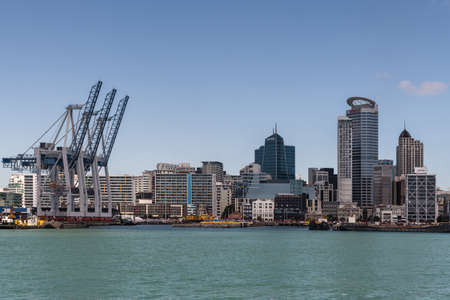 Auckland, New Zealand - March 3, 2017: Row of container cranes at Commercial Harbor with part of city skyline in back under blue sky and behind greenish ocean water. Highrise buildings. No ships.のeditorial素材