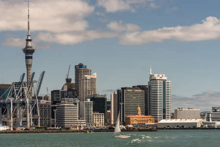 Auckland, New Zealand - March 3, 2017: Container terminal with its cranes under blue sky and behind greenish ocean water. City skyline with ferry terminal and office towers as background. White sailing boat on the water.のeditorial素材