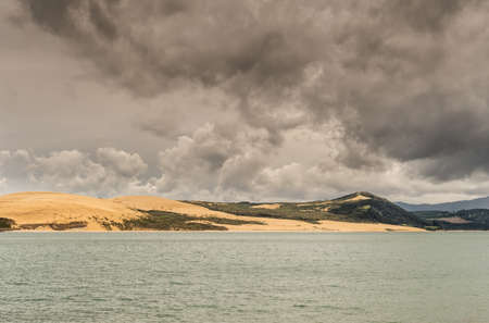 Bay of Islands, New Zealand - March 7, 2017: Exit to Tasman Sea from Hokianga Harbour shows large dune under heavy cloudscape because of approaching cyclone.の写真素材