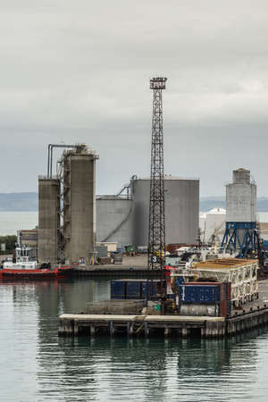 Napier, New Zealand - March 9, 2017: Large fuel tanks and silos at the commercial port under silver sky. Tugboat, light poles, containers and other harbor installations.のeditorial素材