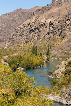 Cromwell, New Zealand - March 15, 2017: Kawarau River meanders and cuts a narrow gorge through dry, brown mountains. Green vegetation borders river. Blue sky.の写真素材