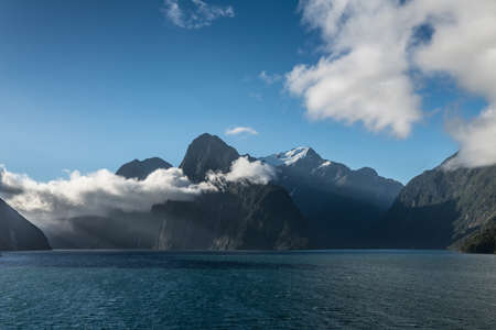 Fiordland National Park, New Zealand - March 16, 2017: Looking over the blue salt waters into the fjord of Milford Sound. Huge mountains, sharp cliffs with low hanging white clouds under blue sky.のeditorial素材