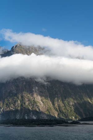 Fiordland National Park, New Zealand - March 16, 2017: Huge cliffs descend into the waters of Milford Sound under blue sky. Green forests at its base. White cloud band hangs midway the rock.のeditorial素材