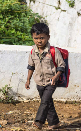 Nilgiri Hills, India - October 26, 2013: A young Tamil schoolboy with beige vest, black pants and red backpack walks to his primary school in the village of Kallatti. Green vegetation and white wall.のeditorial素材
