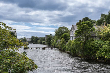 Galway, Ireland - August 3, 2017: The Corrib River rushes between green trees and gray houses to the ocean under heavy cloudscape. Narrow food bridge.のeditorial素材