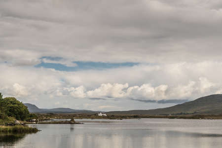 Connemara County, Ireland - August 4, 2017: Wide view of Furnace Lake under a gray sky with one blue patch and pronounced low clouds. Green hills and a white house on the horizon.のeditorial素材