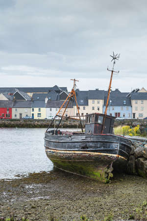 Galway, Ireland - August 3, 2017: The wreck of an old fishing vessel leans against dock wall and stands on dry shore in old Claddagh Port. Colorful houses of The Long Walk Quay form background. Light gray sky.のeditorial素材