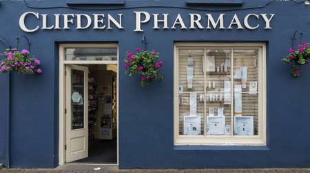 Clifden, Ireland - August 4, 2017: The dark blue facade of the small business pharmacy with white trim, open door and display window. Street scene and flowers on the wall.のeditorial素材