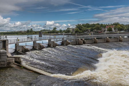 Galway, Ireland - August 5, 2017: Water control dam with open and closed floodgates. Fast flowing white water on Corrib River under blue and white cloudscape. Green trees in background.のeditorial素材