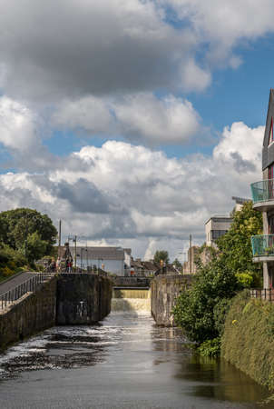 Galway, Ireland - August 5, 2017: Yellow water streams with great force into one side of lock on Eglington Canal downtown under heavy cloudscape. People walking on the shores with trees and houses.のeditorial素材