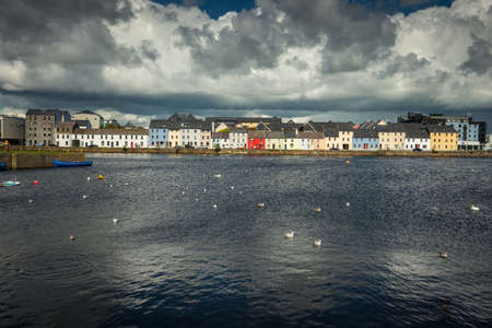 Galway, Ireland - August 5, 2017: Closeup of lIne of colorful houses of The Long Walk quay under enormous black and grey clouds and with dark blue water of the port in front. Some small boats and seagulls.のeditorial素材