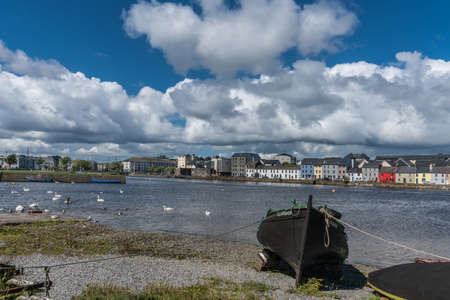Galway, Ireland - August 5, 2017: Enormous white cloudscape in blue sky over section of The Long Walk quay. Up front, black boat on shore. Dark water in between with white swans and seagulls.のeditorial素材