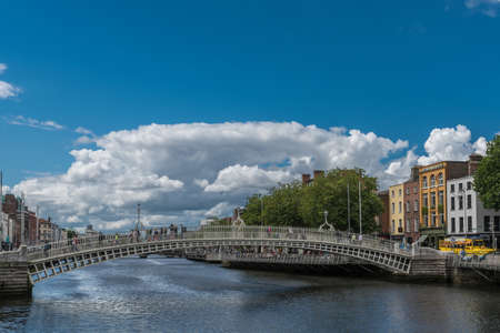 Dublin, Ireland - August 7, 2017: Historic metal Ha'penny pedestrian bridge over Liffey River under blue sky with giant white cloud. People walking. Facades along the river.のeditorial素材