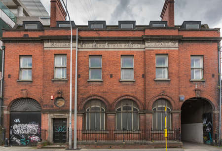 Dublin, Ireland - August 7, 2017: Old dirty abandoned red brick Warehouse along City Quay of Liffey River. Graffiti on gates. Used earlier by British and Irish Steam Packet Company, Ltd. Metal fences.のeditorial素材