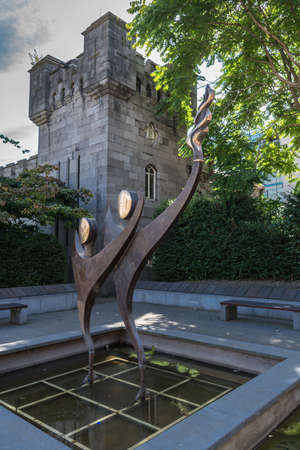 Dublin, Ireland - August 7, 2017: 2003 Special Olympics statue in Dubh-linn garden at The Castle under white sky and green vegetation. Dancing one-legged human figures with flame. Castle wall and tower.のeditorial素材