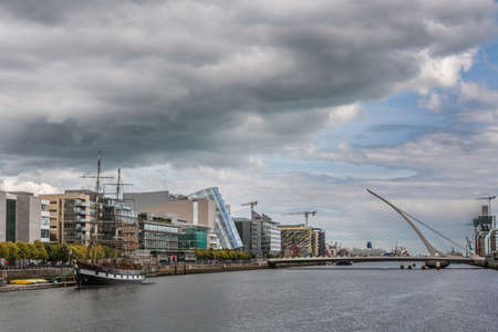 Dublin, Ireland - August 7, 2017: looking east over Liffey River towards modern Samuel Beckett suspension bridge and new Convention Center building under heavy cloudscape. Sail ship.のeditorial素材