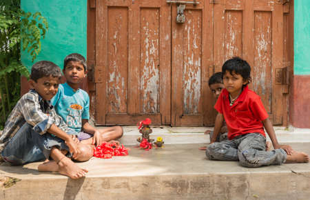 Mysore, India - October 27, 2013: Four small kids in colorful shirts sit and play in front of house with clay figurines looking into camera. Brown and green background.のeditorial素材