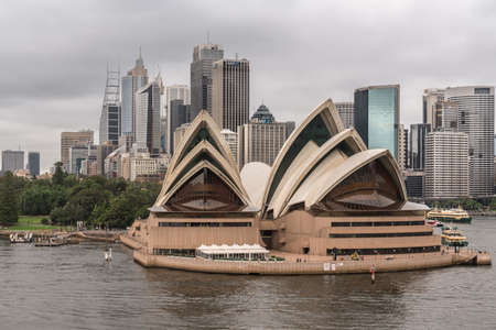 Sydney, Australia - March 21, 2017: Frontal view of Opera House under gray rainy sky. City skyline in back. Water in front, people on the platforms.のeditorial素材