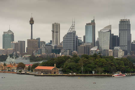 Sydney, Australia - March 21, 2017: Green trees of Garden Island in front of city skyline section with Eye tower under gray sky. Shot from the bay: people on shore line, boats and buoys.のeditorial素材